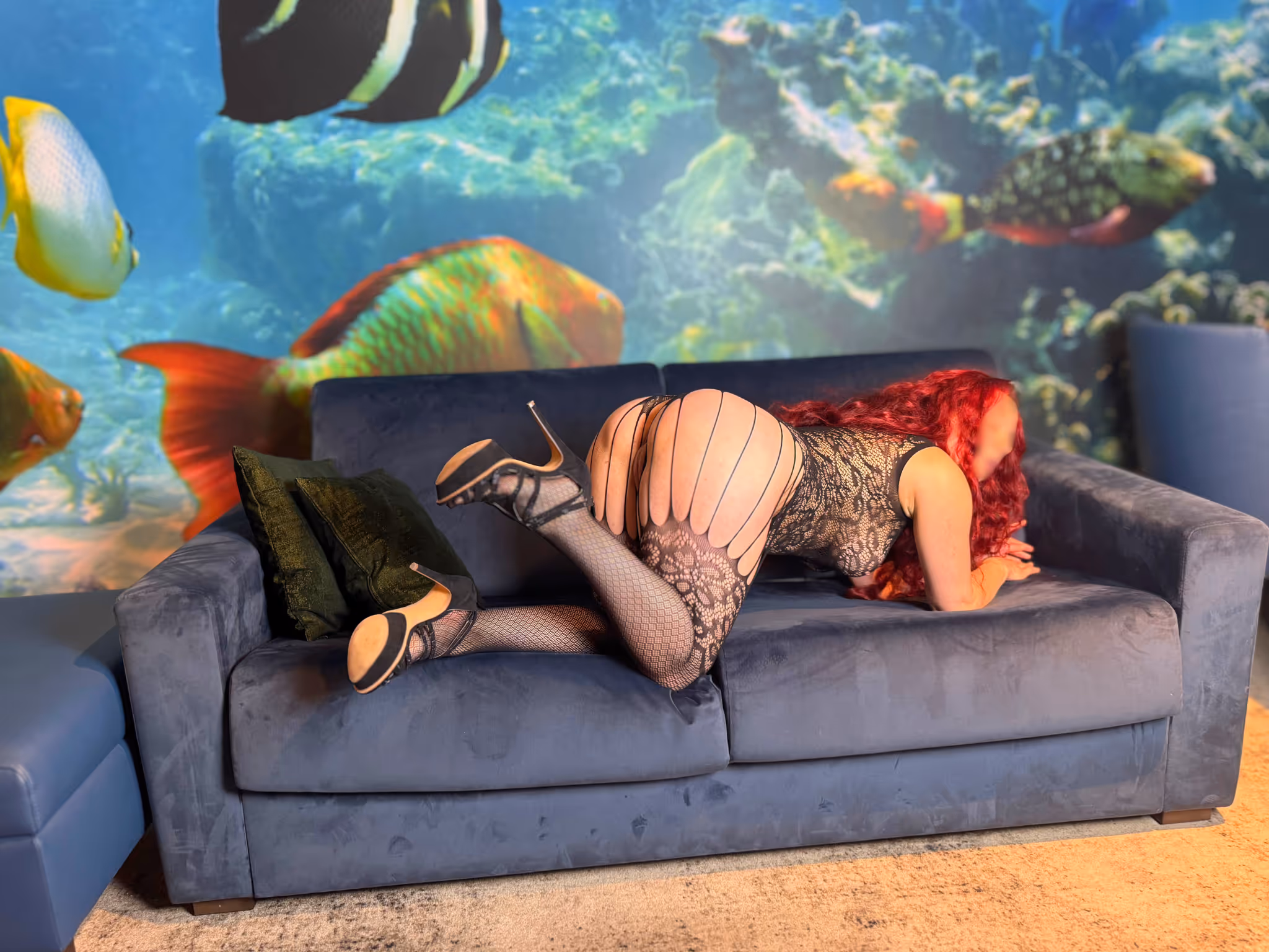 Romy Erfurt Escort liegt entspannt auf dem blauen Samt Sofa in glänzendem rotem Latex Minikleid. Lange rote Haare ausgebreitet schwarze High Heels sie posiert sinnlich mit angehobenem Bein vor buntem Aquarium Wandbild luxuriöses Ambiente Erfurt Escort.